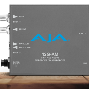 Procesador de audio profesional compacto de color gris con el logotipo de AJA en azul, que muestra indicadores para entrada y salida de vídeo, conexiones ópticas y puerto de audio digital de alta densidad.