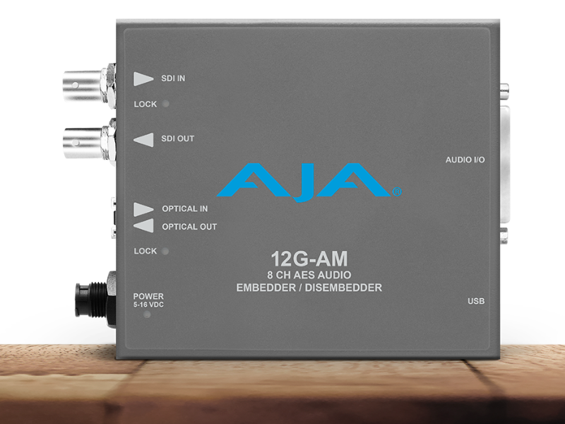Procesador de audio profesional compacto de color gris con el logotipo de AJA en azul, que muestra indicadores para entrada y salida de vídeo, conexiones ópticas y puerto de audio digital de alta densidad.