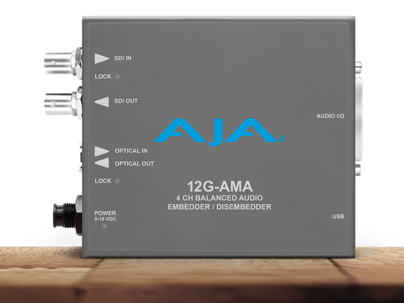 Procesador de audio profesional AJA 12G-AMA en chasis gris, con leyendas blancas que indican la entrada y salida de vídeo SDI, conectividad USB y el puerto de audio balanceado de cuatro canales.