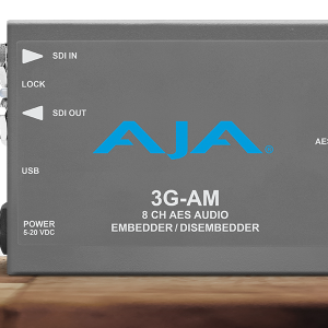 Procesador de audio profesional compacto AJA 3G-AM en chasis gris oscuro, mostrando indicadores para entrada y salida de vídeo SDI, puerto USB y conexión de audio digital de alta densidad.