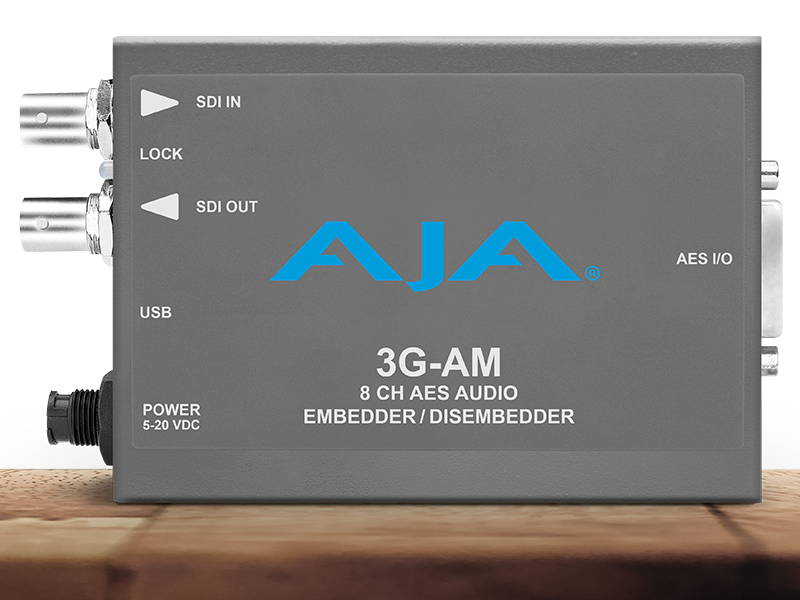 Procesador de audio profesional compacto AJA 3G-AM en chasis gris oscuro, mostrando indicadores para entrada y salida de vídeo SDI, puerto USB y conexión de audio digital de alta densidad.