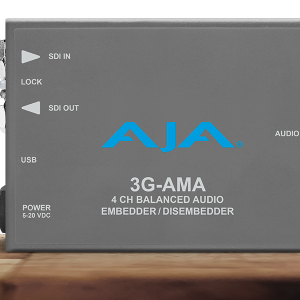 Procesador de audio profesional compacto AJA 3G-AMA en chasis gris oscuro, mostrando los puertos de entrada y salida SDI, el conector USB de configuración y el puerto de audio de alta densidad para señales analógicas balanceadas.