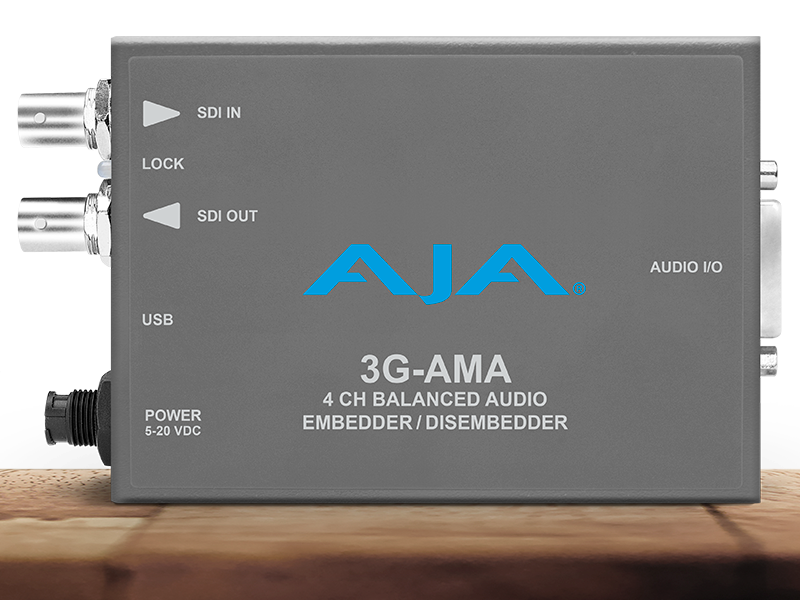 Procesador de audio profesional compacto AJA 3G-AMA en chasis gris oscuro, mostrando los puertos de entrada y salida SDI, el conector USB de configuración y el puerto de audio de alta densidad para señales analógicas balanceadas.