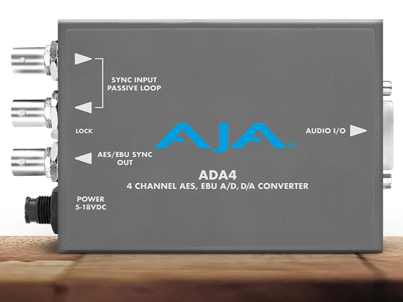 Conversor de audio profesional compacto AJA ADA4 en chasis gris oscuro, mostrando tres conectores BNC para sincronización y salida AES/EBU, puerto USB de configuración y conexión de alimentación circular.