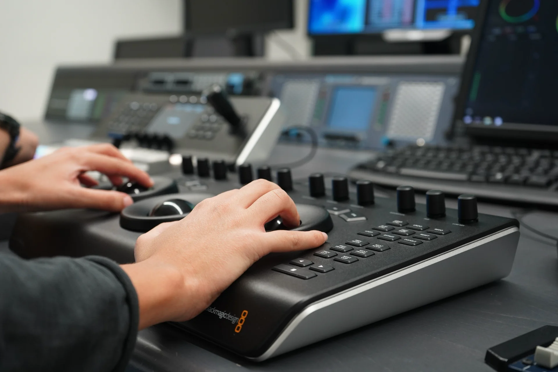 Manos de un técnico operando una consola de gradación de color Blackmagic Design con trackballs y mandos rotativos, con equipos de control y monitores de postproducción desenfocados al fondo